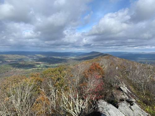 A panoramic view from a mountain peak, showcasing rolling hills, colorful foliage, and a cloudy sky.
