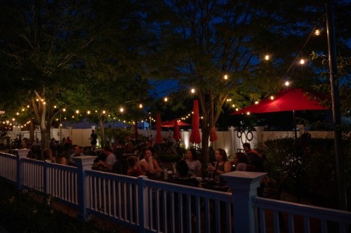 A cozy outdoor restaurant scene at night, with string lights, red umbrellas, and diners enjoying their meals.