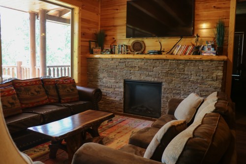 Cozy living room with a stone fireplace, wooden walls, a TV, and comfortable seating.