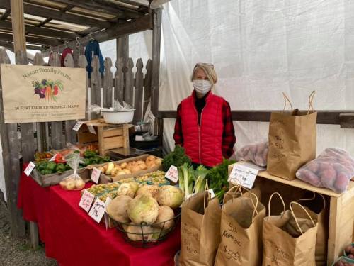 A vendor in a red vest stands at a market stall filled with fresh vegetables and bags of produce.