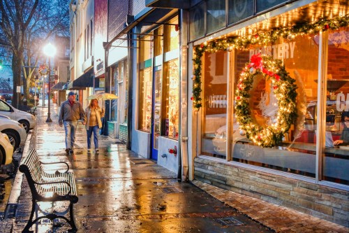 A couple walks under an umbrella past a festive storefront adorned with holiday decorations on a rainy evening.