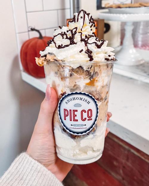 A hand holding a clear cup filled with layers of ice cream, whipped cream, chocolate sprinkles, and pie pieces.