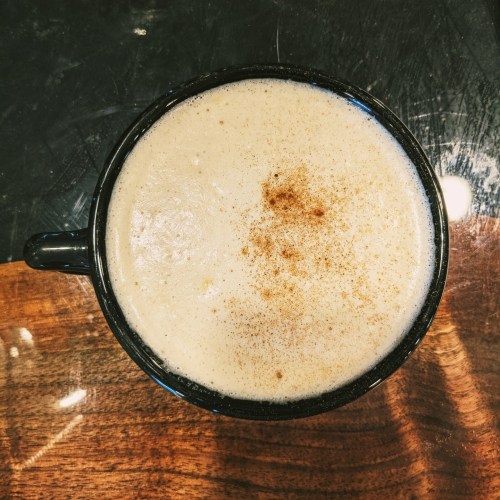 A close-up of a black coffee cup filled with frothy coffee topped with a sprinkle of cinnamon on a wooden table.