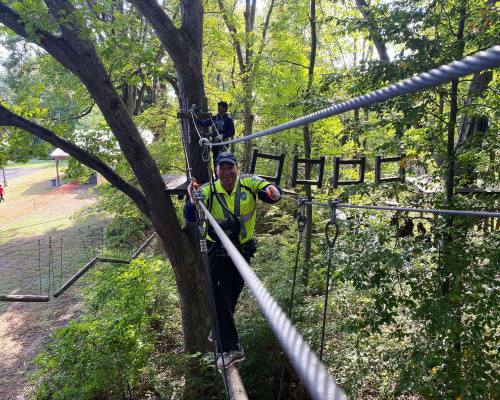 A person in a safety vest balances on a rope bridge among trees, with another person in the background.