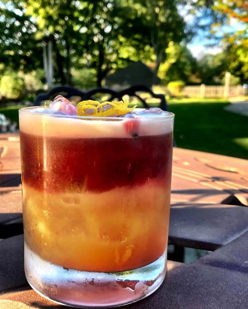 A layered cocktail in a glass, featuring vibrant colors and garnished with citrus, set against a blurred outdoor background.