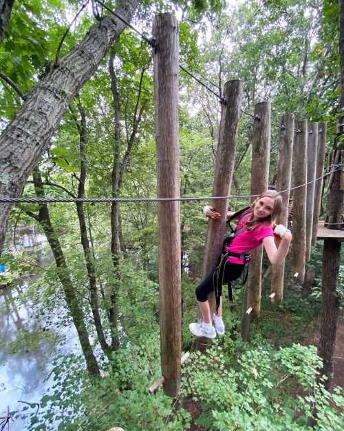 A girl in a pink shirt balances on a rope course among trees, smiling confidently in a lush green setting.