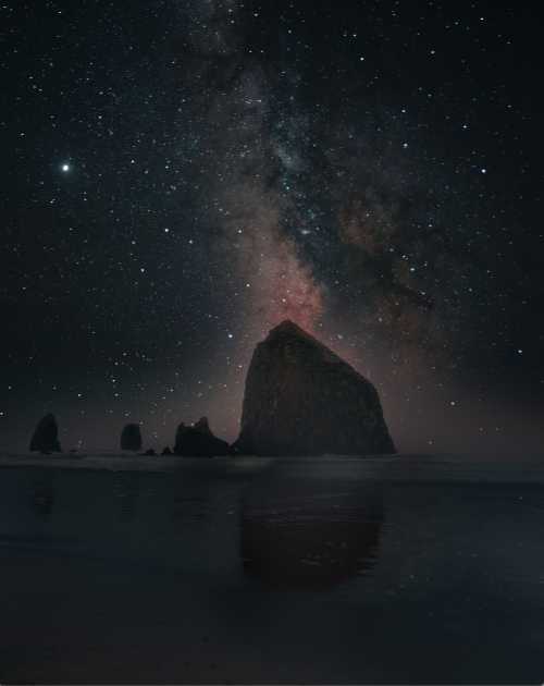 A rocky coastline under a starry night sky, with the Milky Way visible and reflecting on the water.