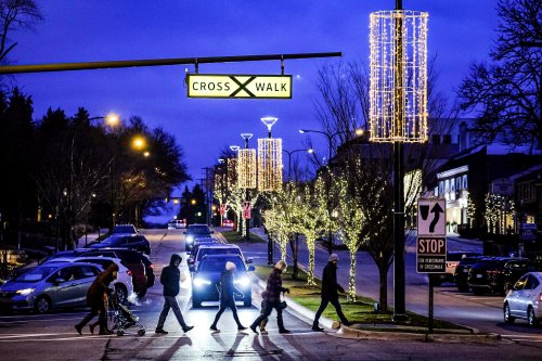 Pedestrians cross a street at dusk, with festive lights and a "CROSS WALK" sign overhead. Cars are parked nearby.