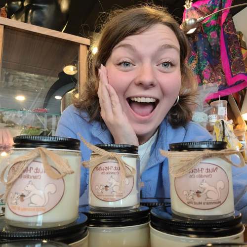 A smiling person poses with jars of candles in a cozy shop, showcasing excitement and joy.