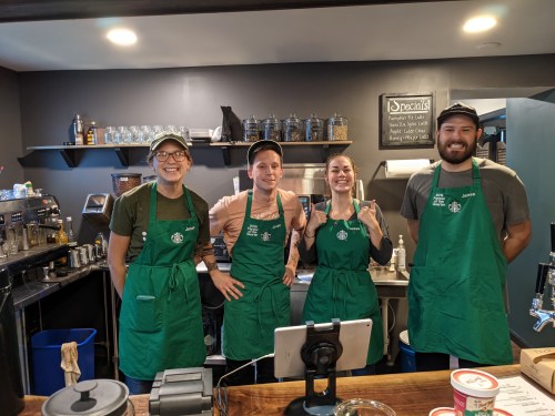 Four baristas in green aprons smile behind a coffee shop counter, with equipment and shelves in the background.