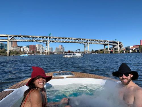 A woman and man relax in a hot tub on a boat, with a city skyline and bridge in the background under a clear blue sky.
