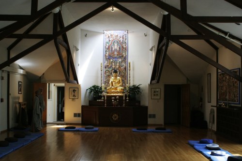 A serene meditation room with a golden Buddha statue, wooden beams, and blue meditation cushions on the floor.
