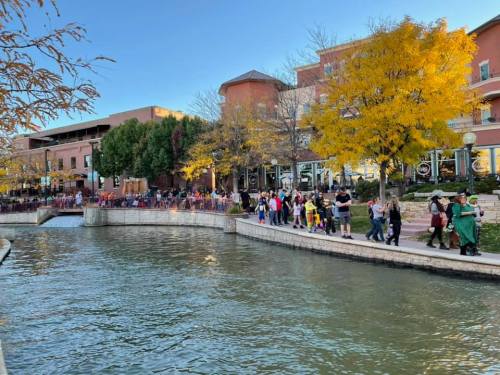 A lively riverside scene with people walking along the bank, colorful trees, and buildings in the background.