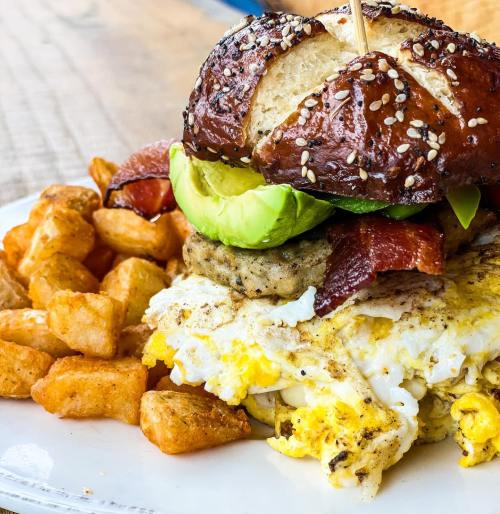 A plate featuring a sesame-seed pretzel sandwich with bacon, avocado, and fried eggs, alongside crispy potato cubes.
