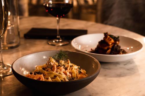 A bowl of pasta garnished with herbs, alongside a plate of dark, textured food and a glass of red wine on a table.