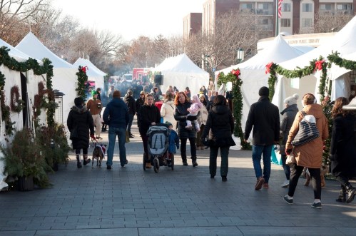 A bustling outdoor market with festive tents, people walking, and holiday decorations in a winter setting.