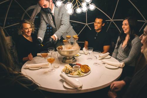 A group of friends enjoys drinks and food at a stylish restaurant, while a waiter serves them at a round table.