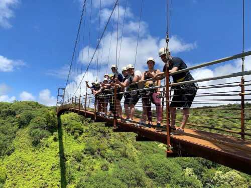 A group of people in helmets stands on a suspension bridge, surrounded by lush green hills and a blue sky.