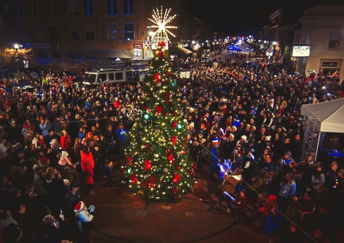 A large crowd gathers around a beautifully lit Christmas tree in a festive night scene.