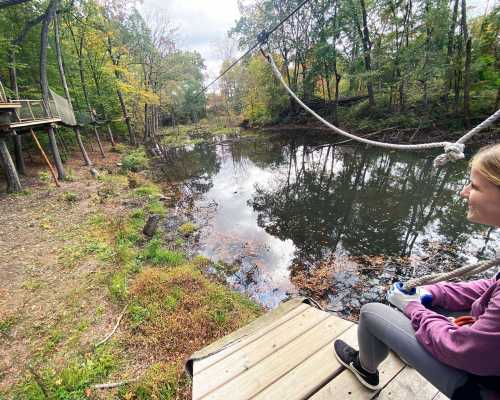 A girl sits on a wooden platform by a calm river, surrounded by trees and autumn foliage.