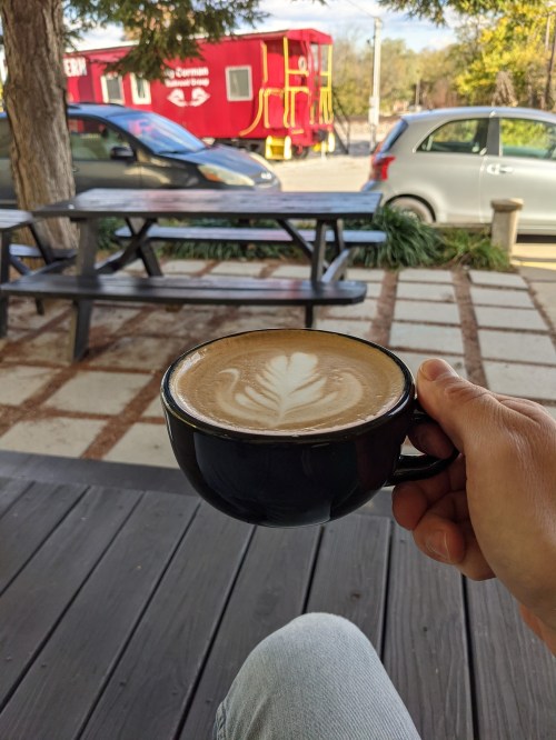 A hand holds a black coffee cup with latte art, seated outdoors with cars and a food truck in the background.