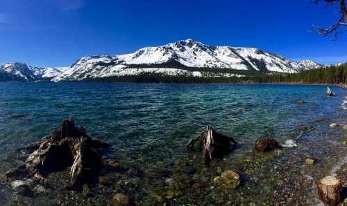 A panoramic view of a clear blue lake surrounded by snow-capped mountains and rocky shores under a bright blue sky.