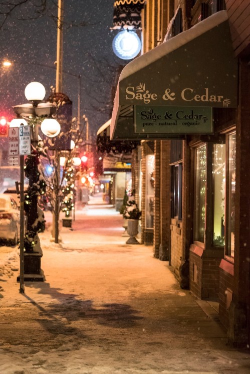 Snowy street scene at night, featuring a shop called "Sage & Cedar" with festive lights and a clock in the background.