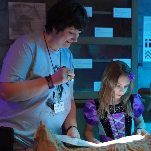 A woman and a girl examine a book together in a cozy, dimly lit room filled with historical displays.