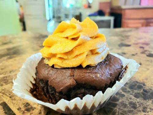 A chocolate cupcake topped with a swirl of orange frosting, placed in a white paper liner on a textured surface.