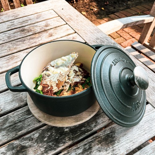 A cast iron pot on a wooden table, filled with food and partially covered by a lid, surrounded by greenery.