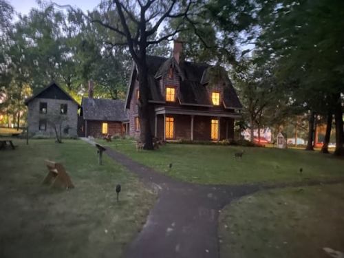A charming house with glowing windows surrounded by trees, set in a serene garden at dusk.