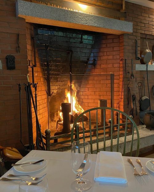 A cozy fireplace with a roaring fire, surrounded by brick walls and dining table settings in a warm atmosphere.