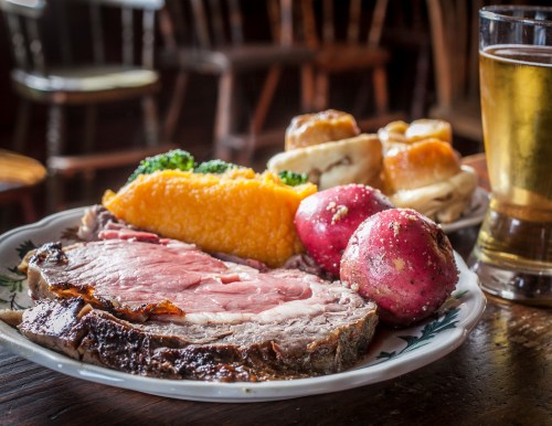 A plate of prime rib with red potatoes, broccoli, and a side of rolls, accompanied by a glass of beer.