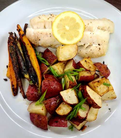 A plate featuring grilled fish with lemon, roasted red potatoes, and colorful carrots, garnished with green onions.