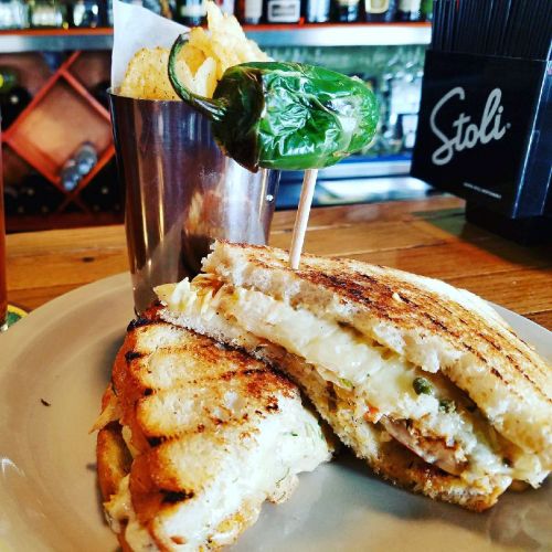 A grilled sandwich with melted cheese and jalapeño, served with a side of chips in a metal cup.