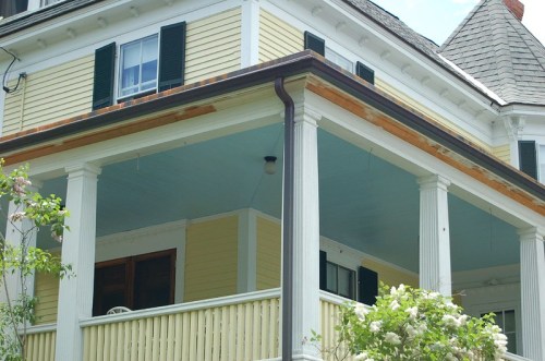 A close-up of a yellow house with a porch, featuring white columns and green shutters, surrounded by greenery.