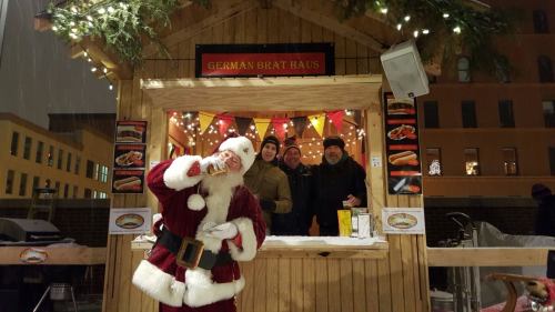 A festive German bratwurst stand with four men, including one in a Santa suit, surrounded by holiday decorations.