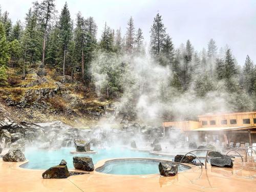 A misty hot spring pool surrounded by rocky terrain and tall trees, creating a serene, natural atmosphere.