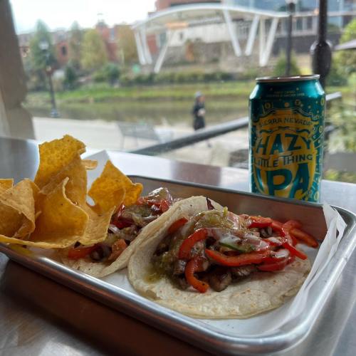 Two tacos filled with meat and vegetables, served with tortilla chips and a can of hazy IPA beer in the background.