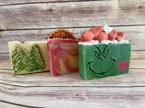 Three colorful handmade soaps: green with a Grinch design, pink swirled, and cream with pine tree patterns, set against a wooden background.