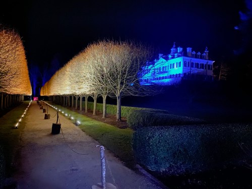 A dimly lit pathway lined with bare trees leads to a blue-lit mansion at night.