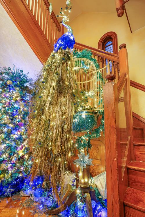A festive staircase adorned with a peacock decoration, twinkling lights, and a decorated Christmas tree nearby.