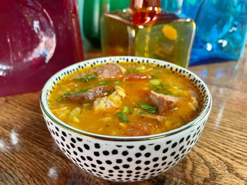 A bowl of hearty soup with meat and herbs, set against colorful glass decorations.