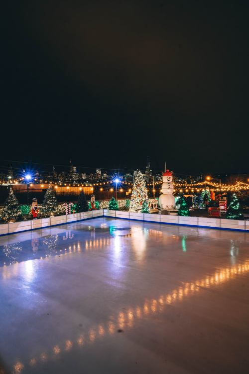 A festive ice skating rink surrounded by decorated Christmas trees and a large snowman, illuminated at night.
