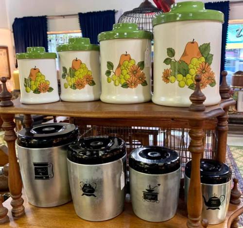 A display of vintage canisters with fruit and floral designs, arranged on a wooden shelf with metal containers below.