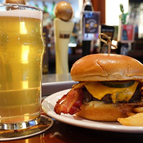 A glass of beer next to a cheeseburger topped with bacon and a pickle, served with fries on a plate.