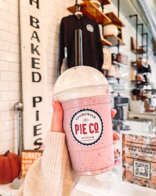 A hand holding a smoothie with whipped cream, featuring a label that reads "Snohomish Pie Co" in a cozy café setting.