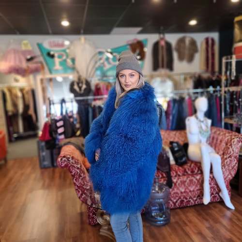 A woman in a blue fur coat and gray beanie poses in a vintage clothing store with colorful decor.