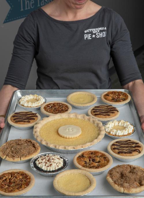 A person holds a tray filled with various pies, including pecan, chocolate, and lemon, showcasing a variety of flavors.