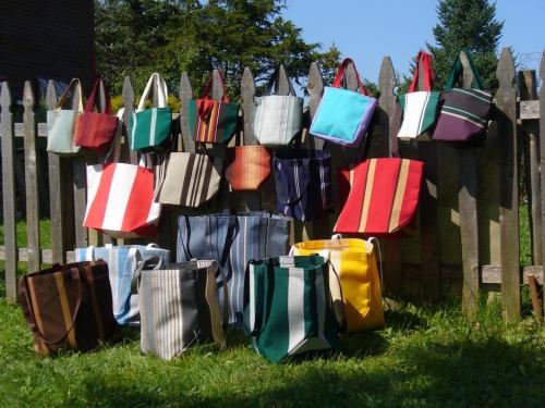A variety of colorful tote bags hanging on a wooden fence, set against a sunny outdoor background.